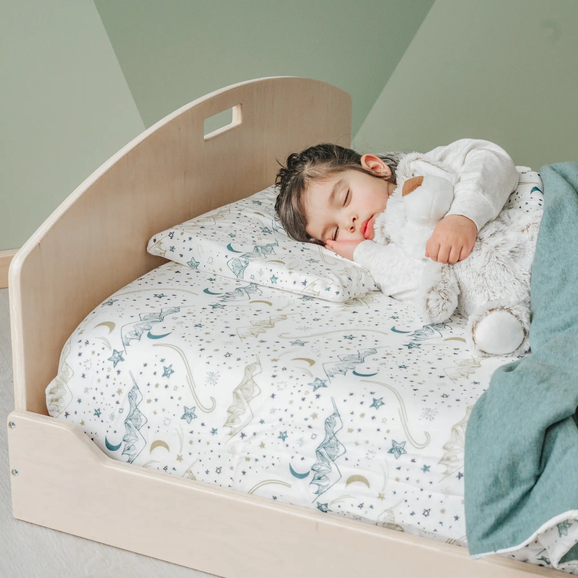 Toddler sleeping on a low wooden bed with a curved headboard, designed for safe, independent sleep. The bed has celestial-patterned bedding with stars and moons. The child, in a white long-sleeve outfit, hugs a plush toy while resting. A sage green blanket adds warmth. The natural wood finish pairs with the muted green wall for a Montessori-inspired look.