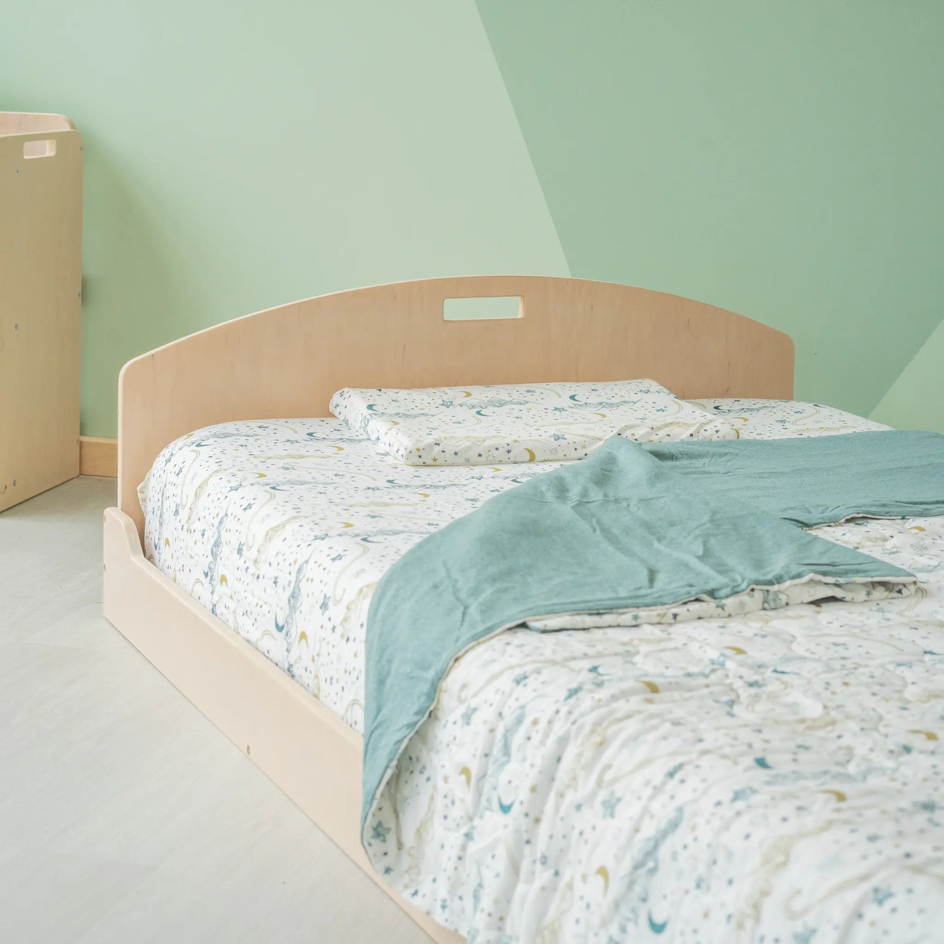 Low wooden bed with a curved headboard, dressed in celestial-patterned bedding with stars and moons. A soft sage green blanket adds warmth. The Montessori-inspired bedroom features muted green walls and light wood flooring, creating a calming sleep space.
