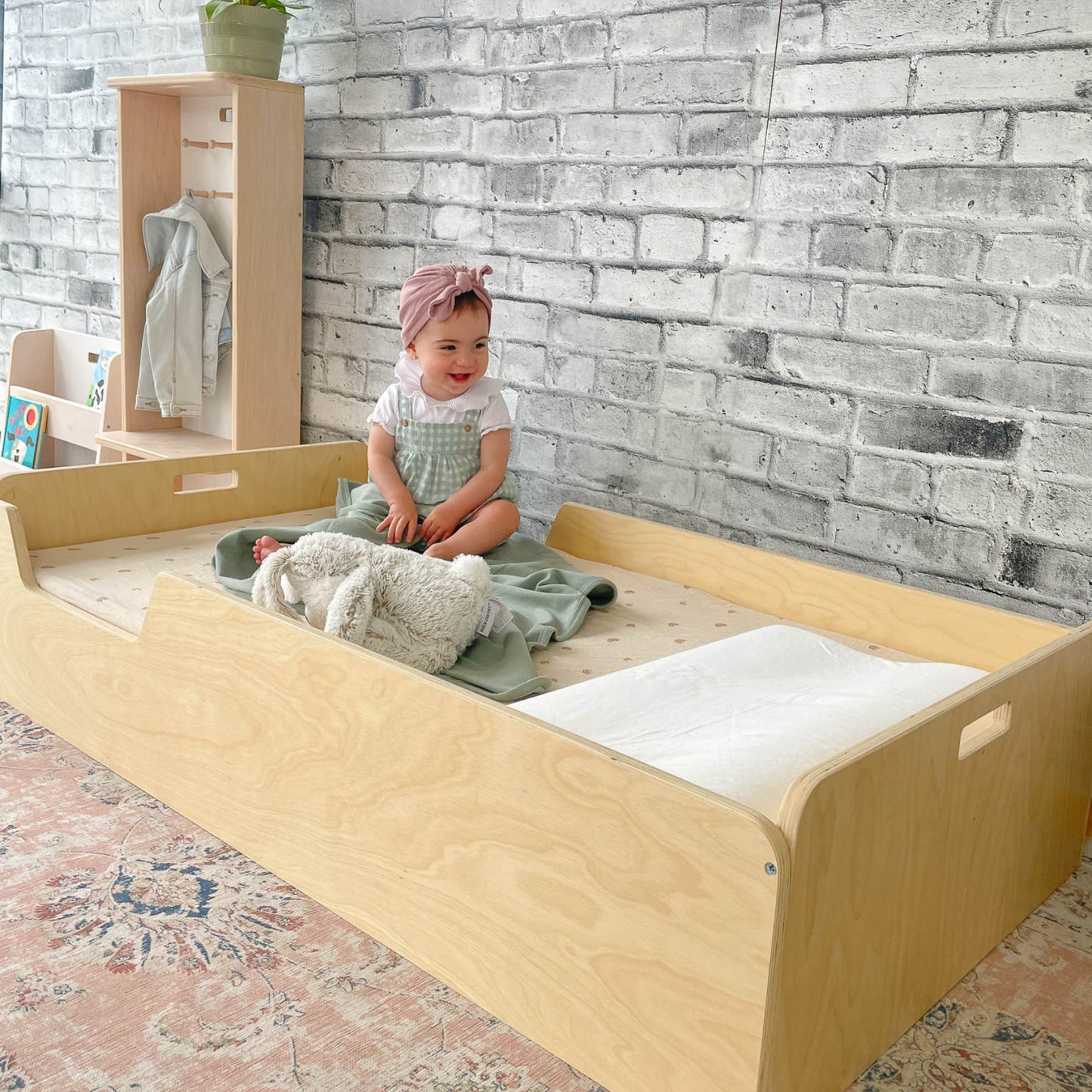 Baby smiling and sitting on a low enclosed wooden bed, designed for independent sleep. The Montessori-style bed features built-in handles and a perforated base. A plush toy, sage green blanket, and open shelving create a cozy toddler space.