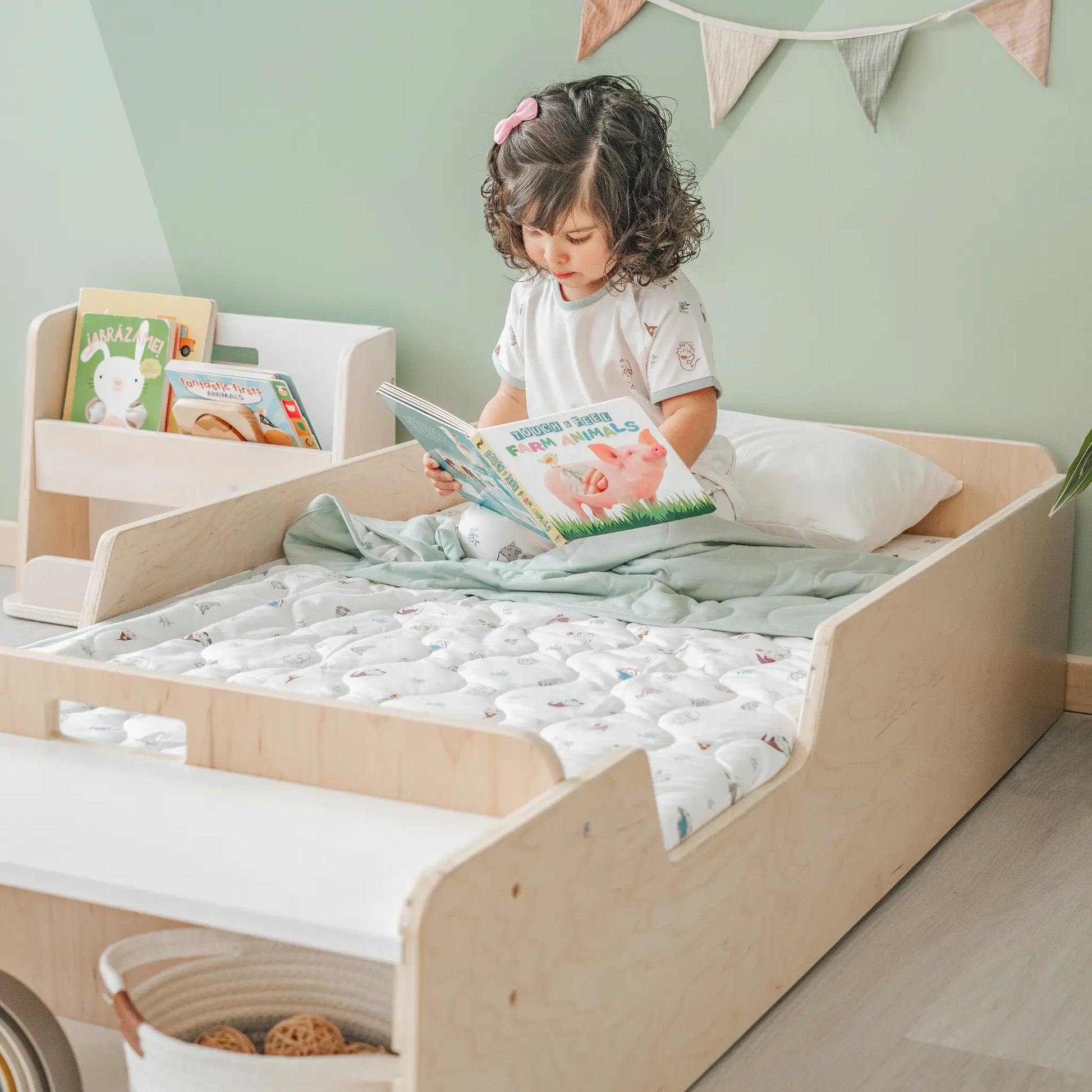 Young girl reading a book on a Montessori junior floor bed made of birch wood, featuring front storage shelf and toddler-safe low profile design – styled in a serene Montessori bedroom with soft green walls and wooden book display – encourages independent reading and calm routines – Sapiens Child bed, handcrafted in Canada, available in birch or maple
