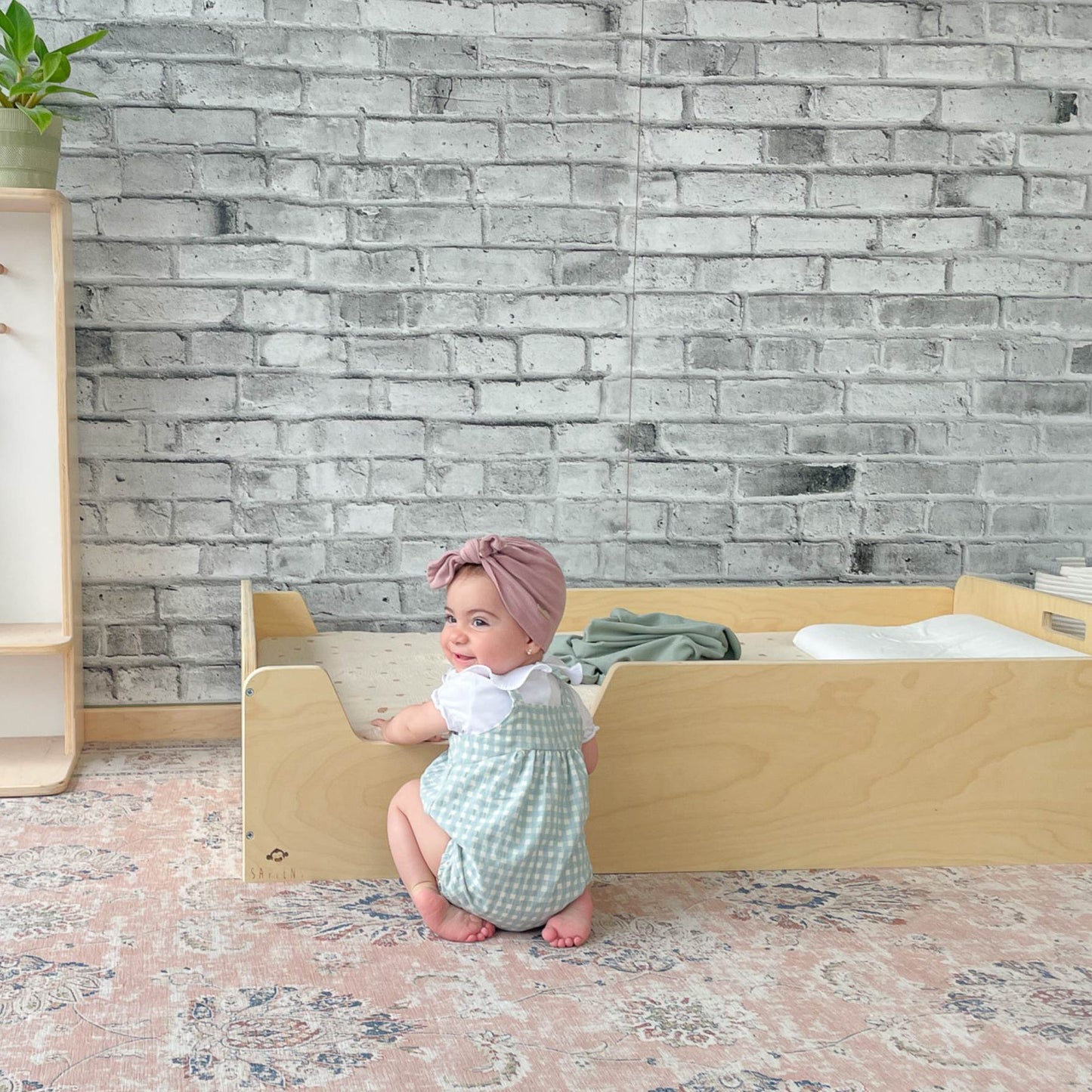 Baby standing beside a low enclosed wooden bed, designed for independent sleep. The Montessori-style bed allows easy access for toddlers. A soft rug, brick wall, and open shelving create a cozy, child-friendly space.