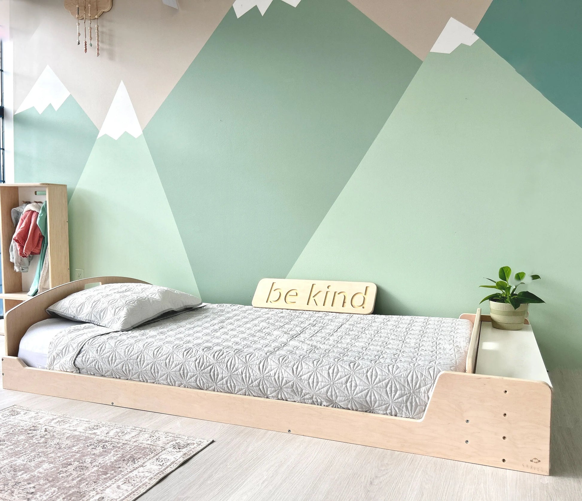 Low wooden floor bed with built-in side storage, styled in a cozy Montessori-inspired bedroom. The minimalist design supports independent sleep while keeping essentials within reach. A soft gray quilt, neutral decor, and a mountain mural create a calming, nature-inspired space.