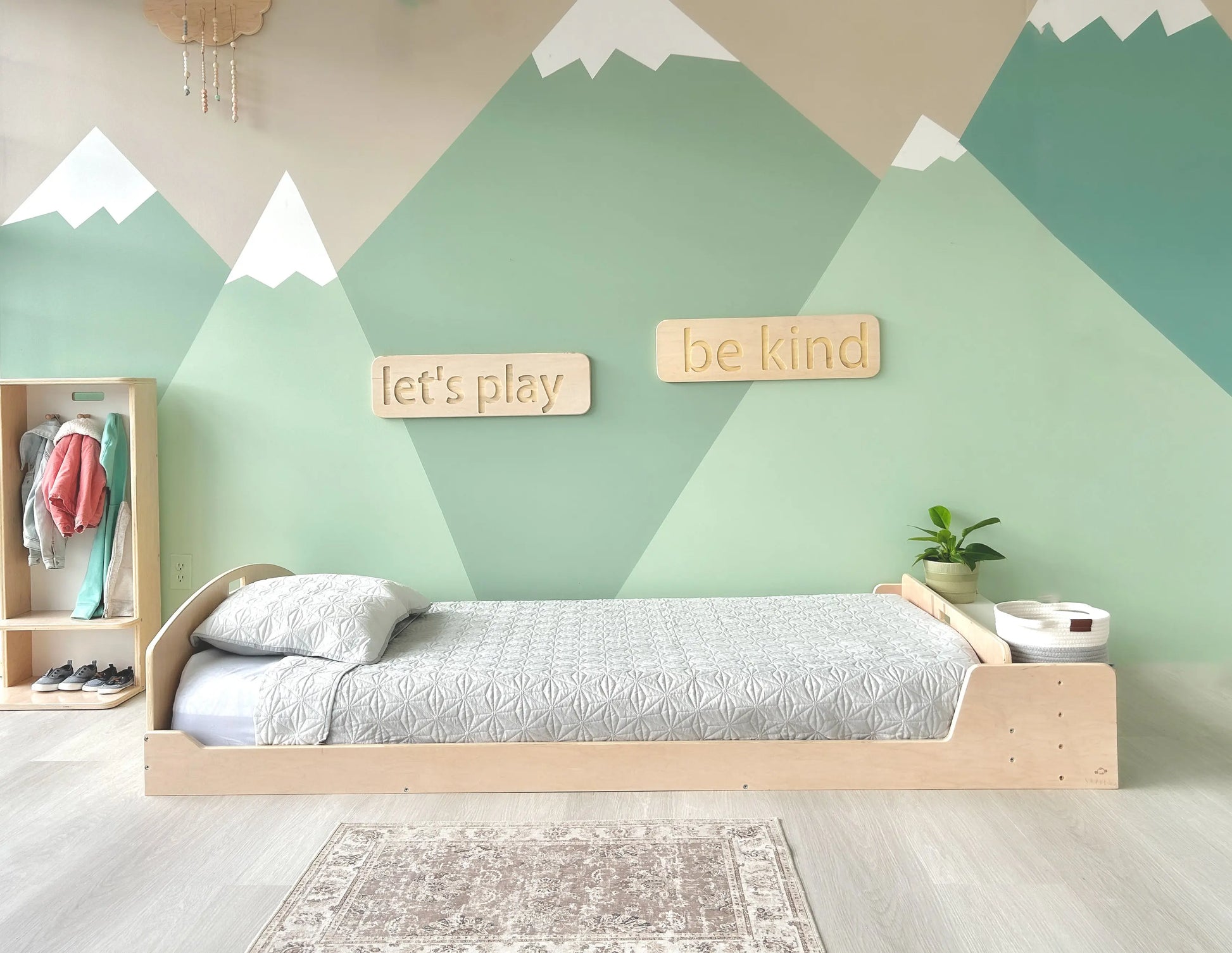 Low Montessori-style floor bed with built-in storage, set in a calming nature-inspired kids' room. A mountain mural, soft gray bedding, and wooden wall decor reading "let’s play" and "be kind" create a playful yet serene space. Designed for independent sleep and organization, the bed blends functionality with minimalist aesthetics.