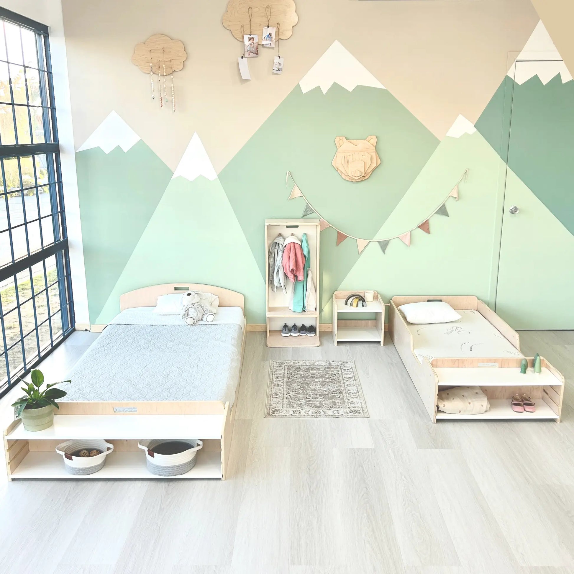 Montessori bedroom setup featuring two low wooden beds with customizable storage shelves at the foot. The adjustable shelves can hold books, slippers, plants, or a water cup, adapting to a child’s needs. A coordinated clothing rack and bookshelf encourage independence, while the mountain mural and natural light create a peaceful, organized space.
