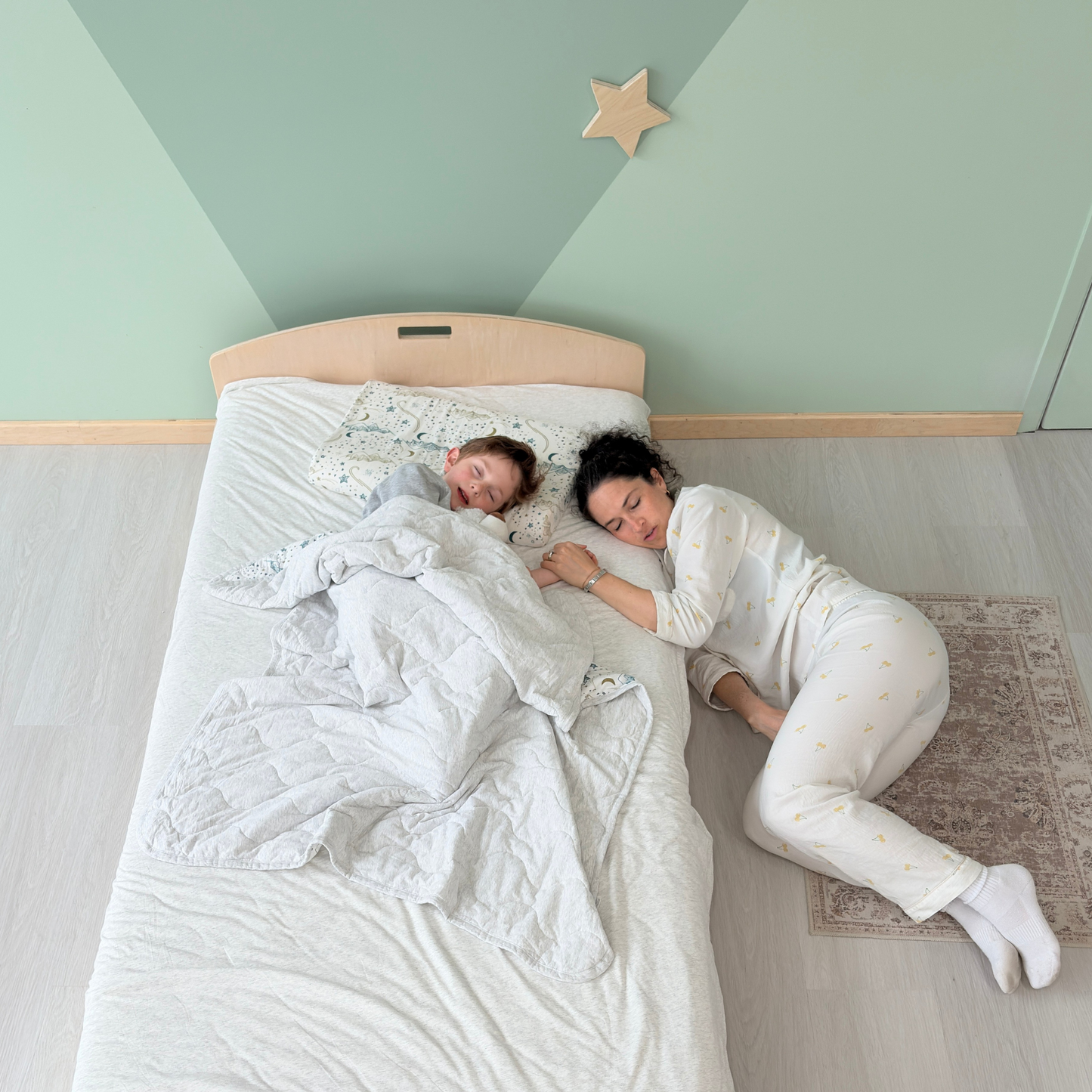 Parent resting on the floor beside a peacefully sleeping child, gently holding hands across the edge of a low wooden bed—capturing the quiet presence and emotional closeness this bedtime setup allows