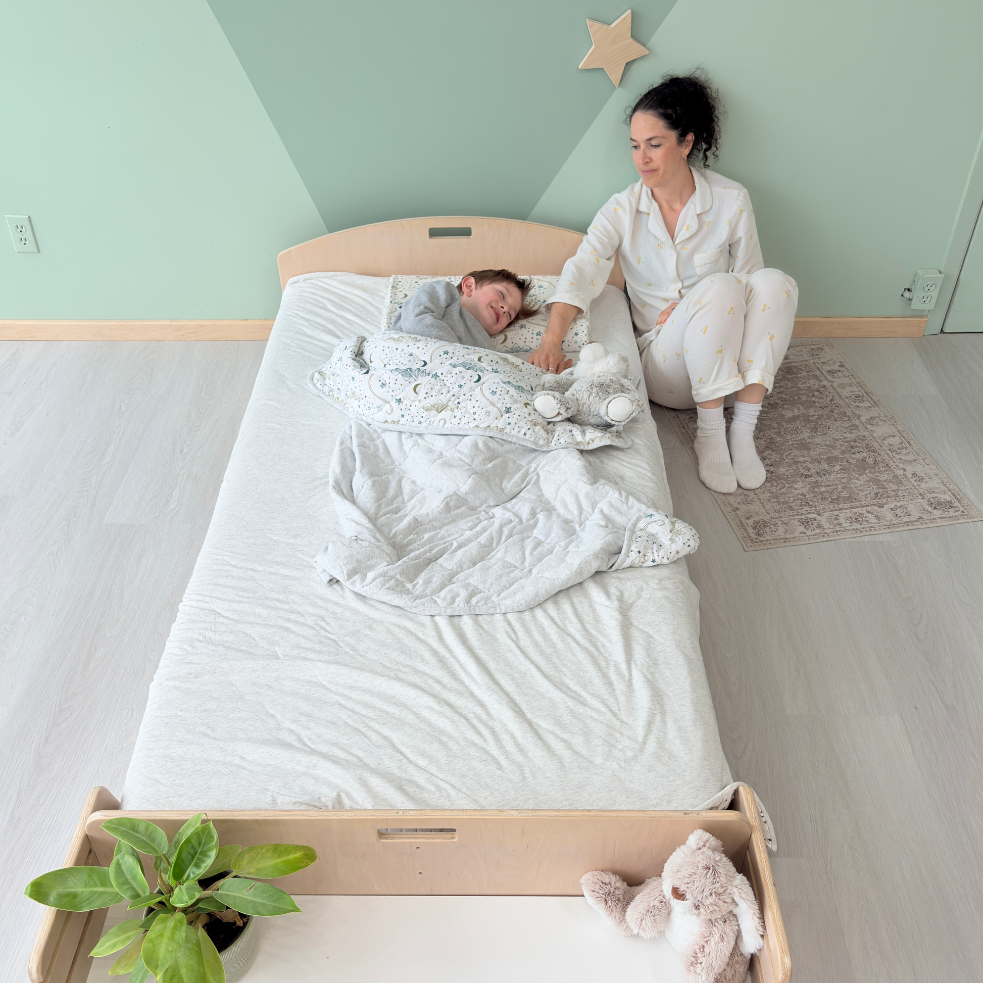 Mother sitting beside her sleeping child on a Montessori twin floor bed made of birch wood, gently offering comfort during bedtime – features low-profile design with built-in shelf, toddler-friendly access, and cozy bedding – ideal for Montessori sleep routines, emotional security, and gentle nighttime transitions – handcrafted in Canada by Sapiens Child, available in birch or maple