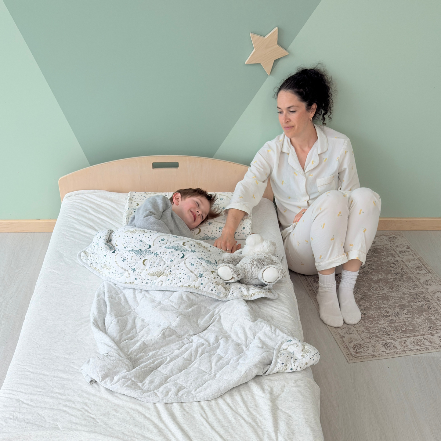 Mom sitting quietly beside her child during bedtime, offering gentle reassurance while the child rests peacefully on a low wooden bed with rounded edges and cozy bedding