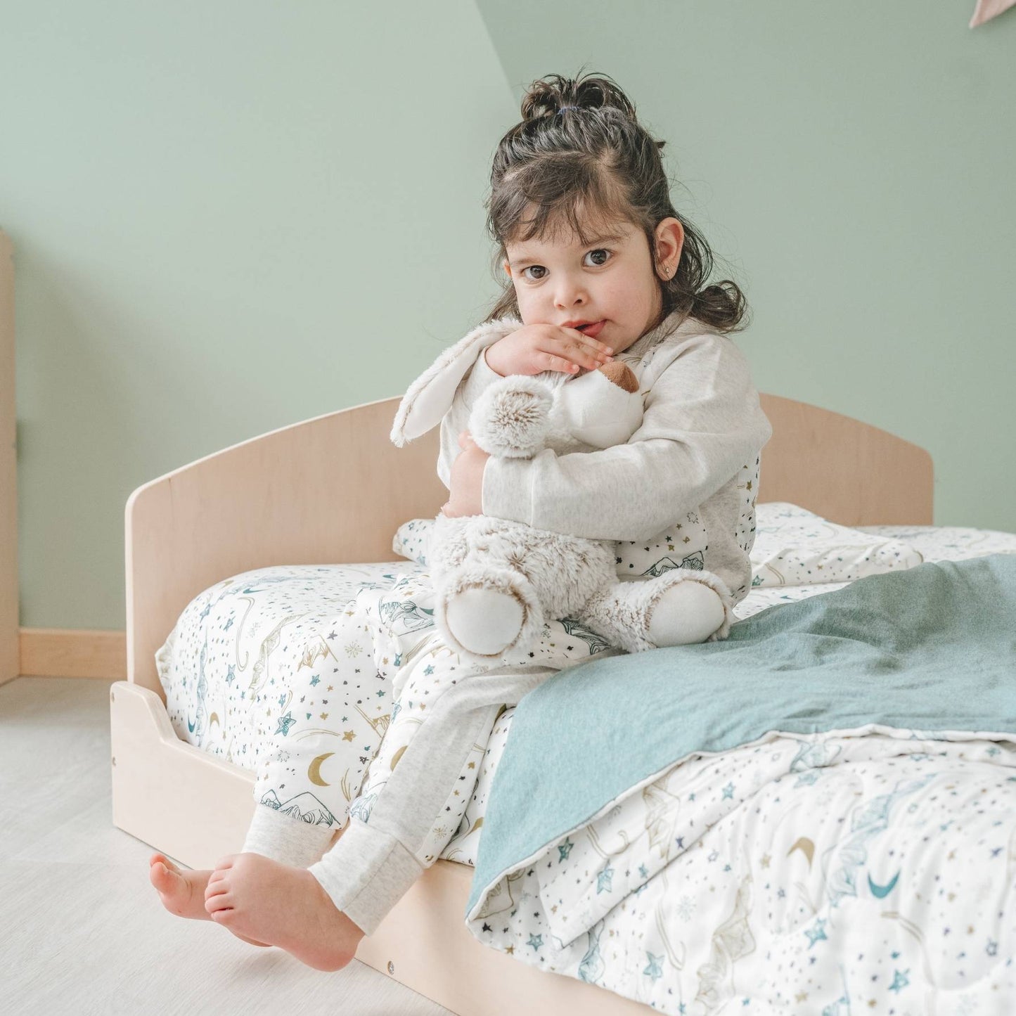 Toddler sitting on a low wooden bed, hugging a plush bunny. The bed features a curved headboard and celestial-patterned bedding with stars and moons. A sage green blanket adds warmth. The child, dressed in soft pajamas, gazes at the camera. A Montessori-inspired room with muted green walls creates a calming space.