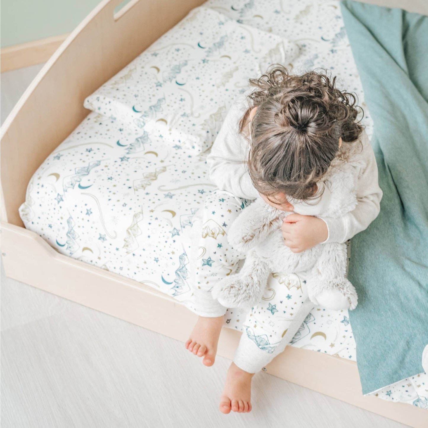 Toddler sitting on a low wooden bed, feet touching the floor for easy access. Designed for safe, independent sleep, this floor-level bed allows young children to climb in and out without assistance. The celestial-themed bedding and sage green blanket create a cozy, Montessori-inspired space.