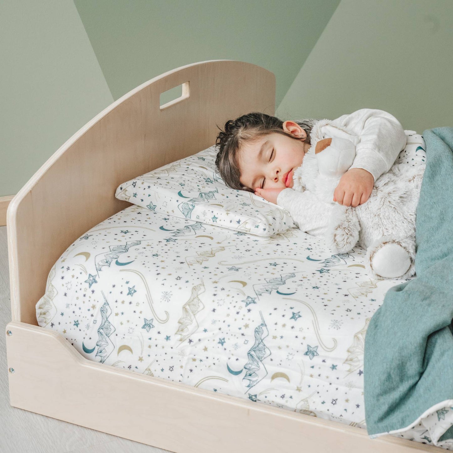 Toddler sleeping on a low wooden bed with a curved headboard, designed for safe, independent sleep. The bed has celestial-patterned bedding with stars and moons. The child, in a white long-sleeve outfit, hugs a plush toy while resting. A sage green blanket adds warmth. The natural wood finish pairs with the muted green wall for a Montessori-inspired look.