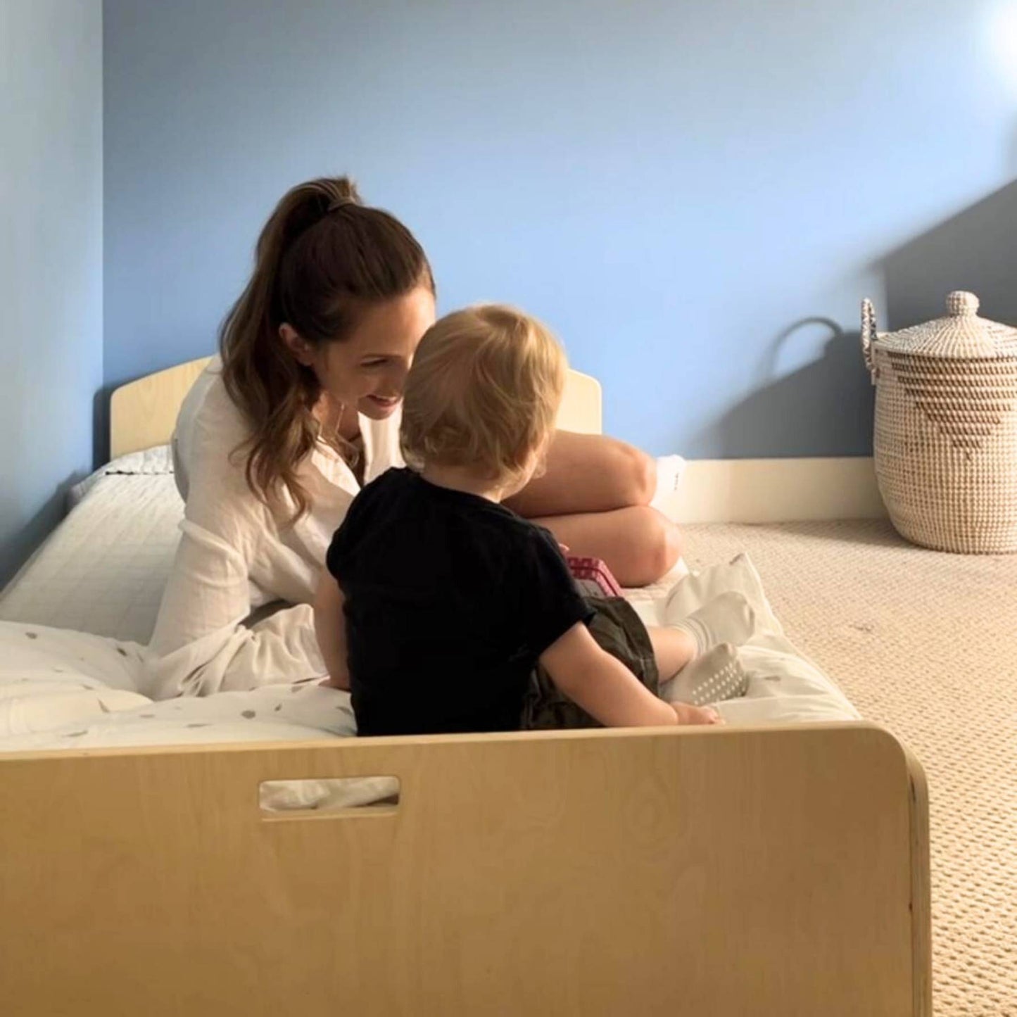 Mother and toddler sitting on a low wooden bed, sharing a moment. The bed has a natural wood finish, curved edges, and white bedding. A blue wall and woven basket add to the cozy, Montessori-inspired room. Safe, accessible sleep space for young children.