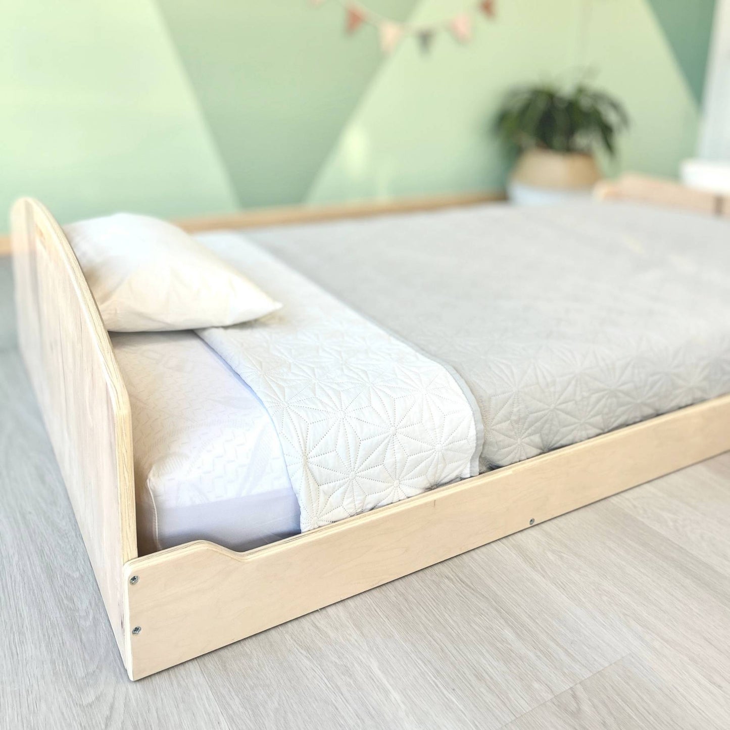 Low wooden bed with a minimalist design, ideal for safe, independent sleep. The floor-level frame allows toddlers easy access while reducing fall risks. Dressed in soft gray and white bedding, the setup creates a calming, Montessori-inspired sleep space. Light wood flooring and a muted green accent wall enhance the natural aesthetic.