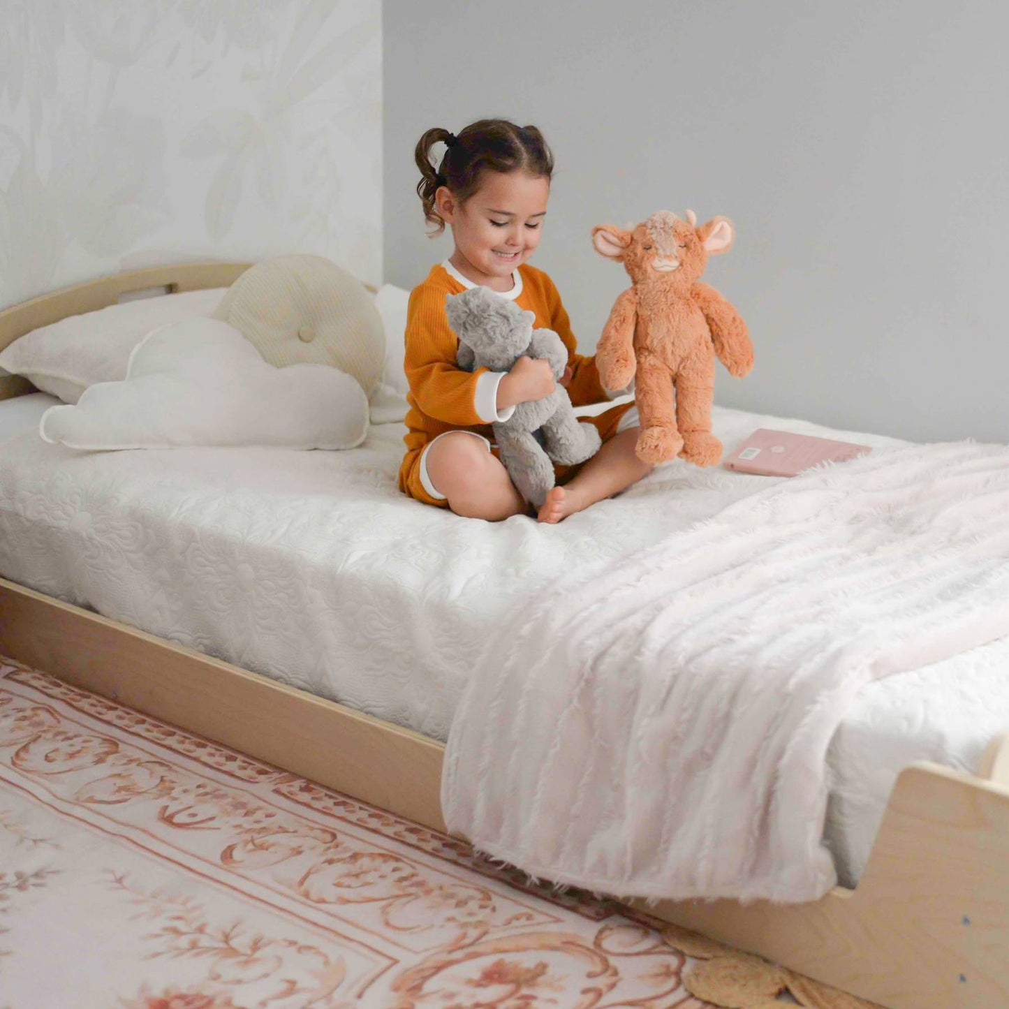 A toddler girl sitting on a low-profile wooden bed, designed for independent sleep and safe transitions from a crib. The bed features a minimalist design with rounded edges and a sturdy frame. It is styled with a white textured quilt, a soft faux-fur throw blanket, and cloud-shaped pillows for a cozy and inviting atmosphere. The child, dressed in a mustard-colored pajama set, is happily playing with two plush stuffed animals. A vintage-inspired area rug in warm beige and brown tones adds to the soothing, Mo