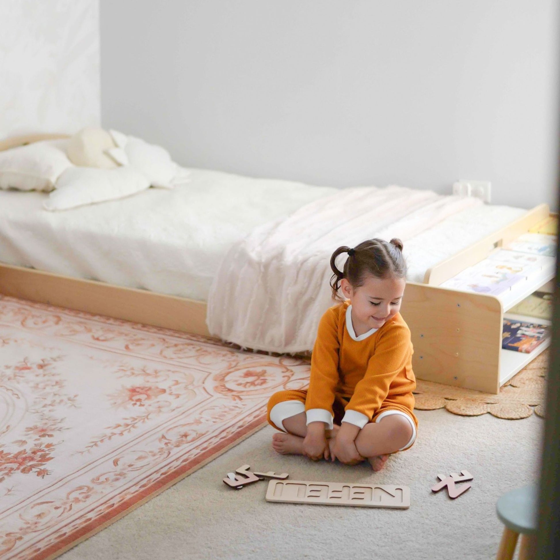 Cozy Montessori-inspired kids' bedroom featuring a natural wood floor bed with an integrated bookshelf. A young child in a mustard-colored pajama sits on the floor, playing with a wooden name puzzle. The space is designed with neutral tones, a vintage-patterned rug, and minimalistic decor to encourage independence and learning.