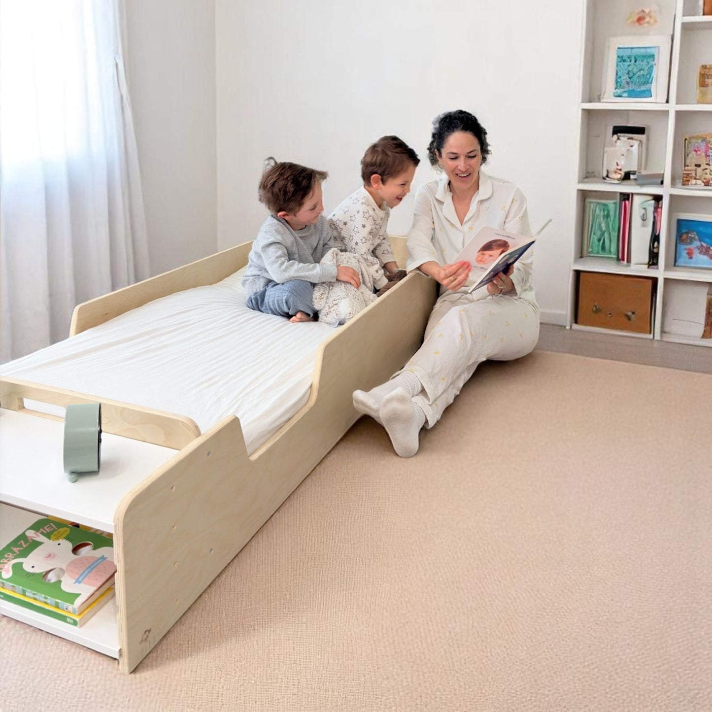 Mother reading a bedtime story to two young children seated on a Montessori junior floor bed in natural birch wood, with integrated shelf holding children’s books – cozy minimalist nursery setup promoting bonding, independence, and calm bedtime routines – safe toddler bed alternative, handcrafted in Canada by Sapiens Child, available in birch or maple wood