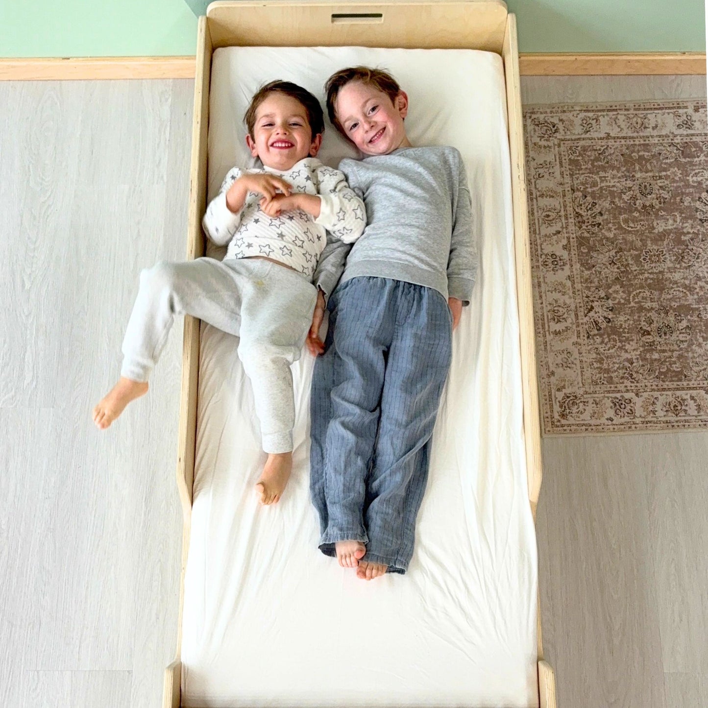 Two smiling children, ages 3 and 6, lying side by side on a Montessori junior floor bed made of birch wood – demonstrates ample space for growing kids and sibling bonding – low-to-ground design with soft edges, ideal for Montessori-inspired bedrooms and independent sleep – handcrafted in Canada by Sapiens Child, available in birch or maple wood