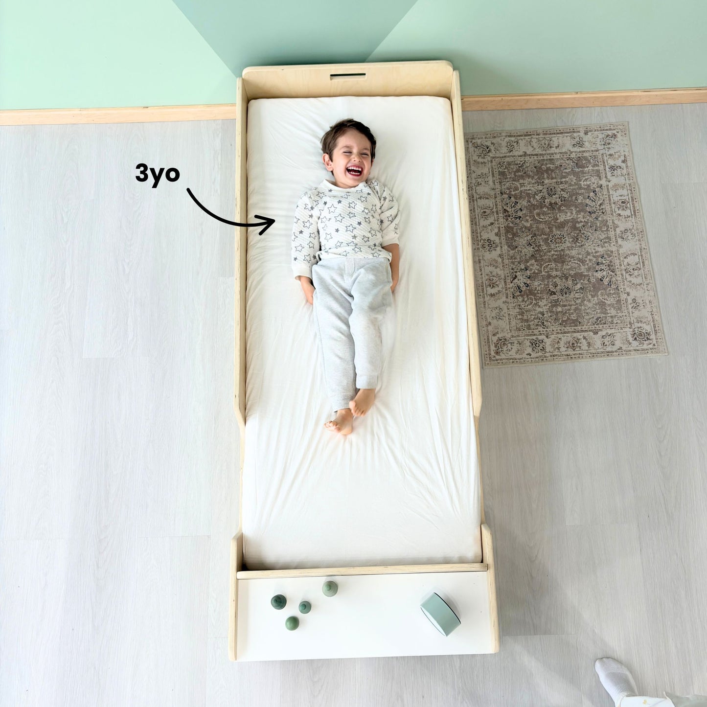 Top view of a happy 3-year-old child lying on a Montessori junior floor bed made of birch wood, with plenty of room to grow – low-profile design with built-in front shelf, safe for toddler transitions from crib to bed – perfect for Montessori-inspired bedrooms and independent sleep routines – handcrafted in Canada by Sapiens Child, available in birch or maple