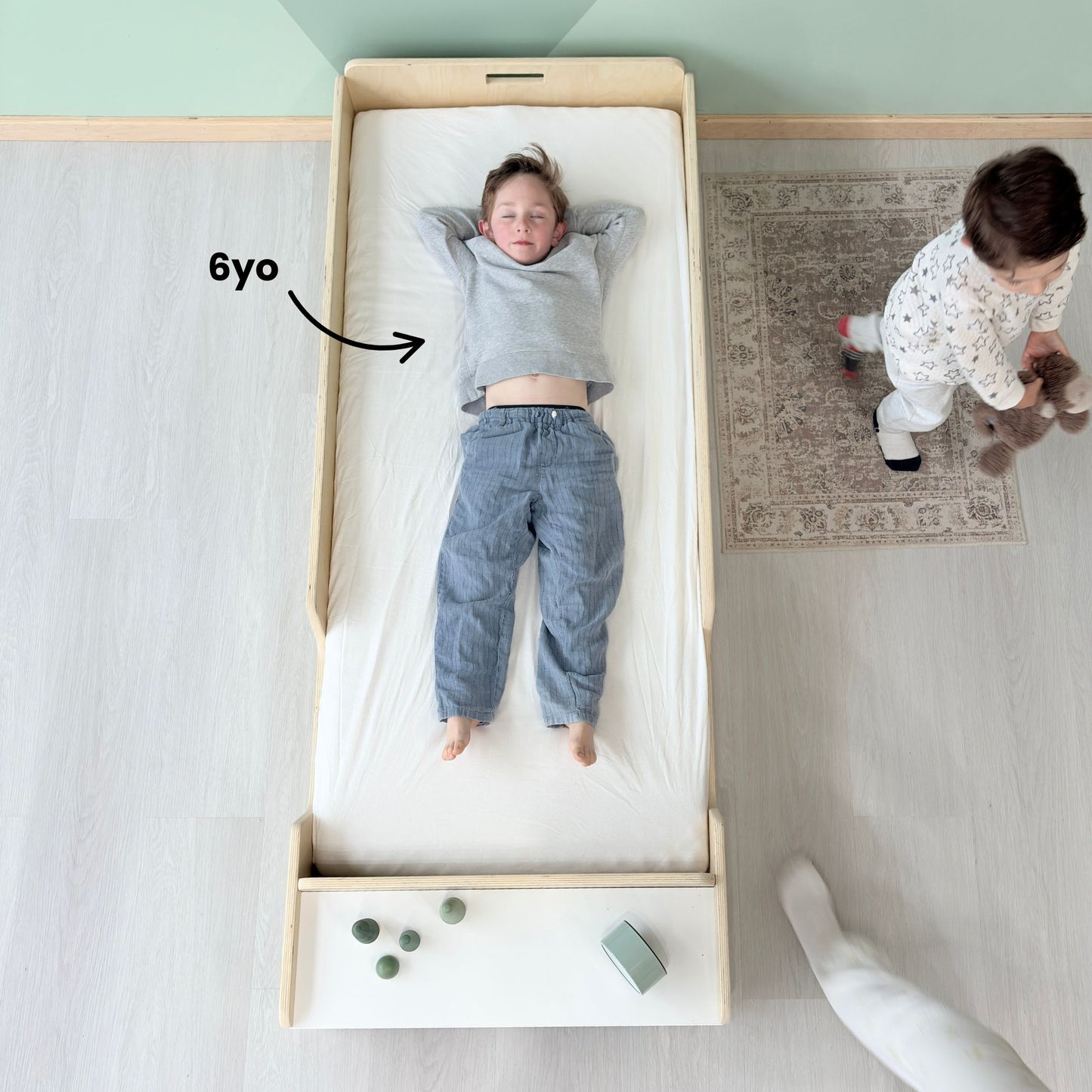 Top view of a 6-year-old child lying comfortably on a Montessori junior floor bed in birch wood, with room to grow – shown with built-in front shelf and minimalist Scandinavian-style kids room – demonstrates spacious fit and extended usability for older toddlers and young children – handcrafted in Canada by Sapiens Child, available in birch or maple wood