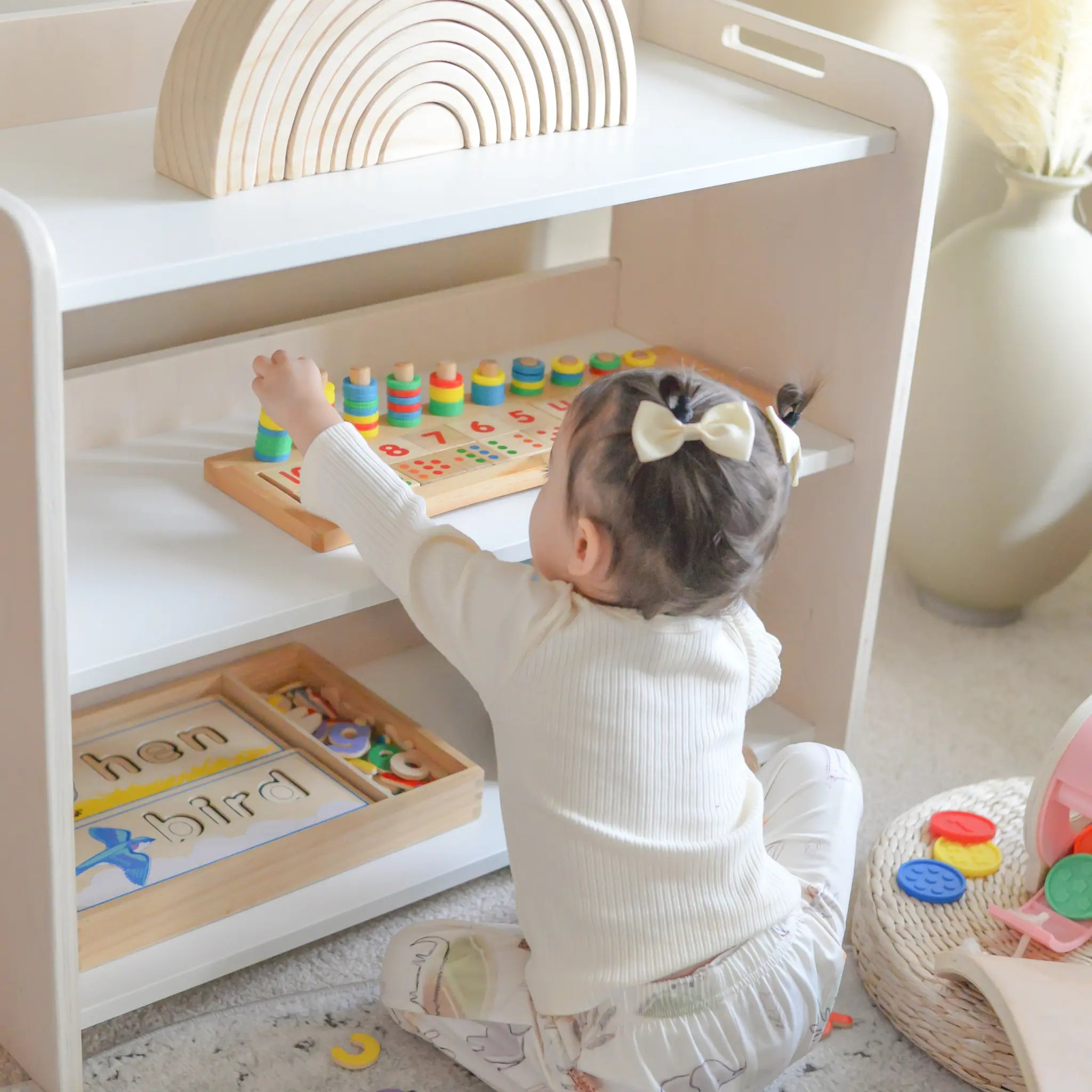Young toddler independently playing with Montessori toys on a low birch shelf, designed for easy access. The child reaches for a colorful counting puzzle, surrounded by natural wooden learning materials, demonstrating the shelf’s child-led, organized setup in a cozy, light-filled space, from Sapiens Child.