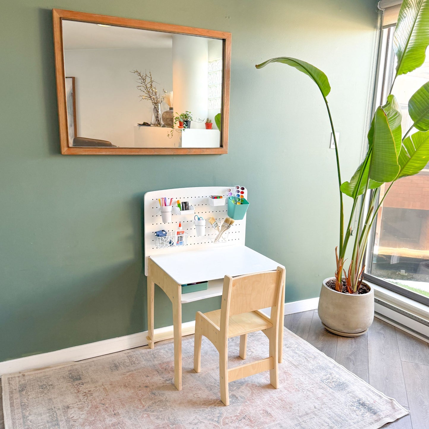 A modern, minimalist children's desk and chair set made of natural wood, featuring an adjustable pegboard organizer with various art supplies. The desk is positioned against a green accent wall with a large mirror above it, and a tall potted plant nearby, creating a cozy and inviting workspace for kids. The setup is illuminated by natural light from a large window.