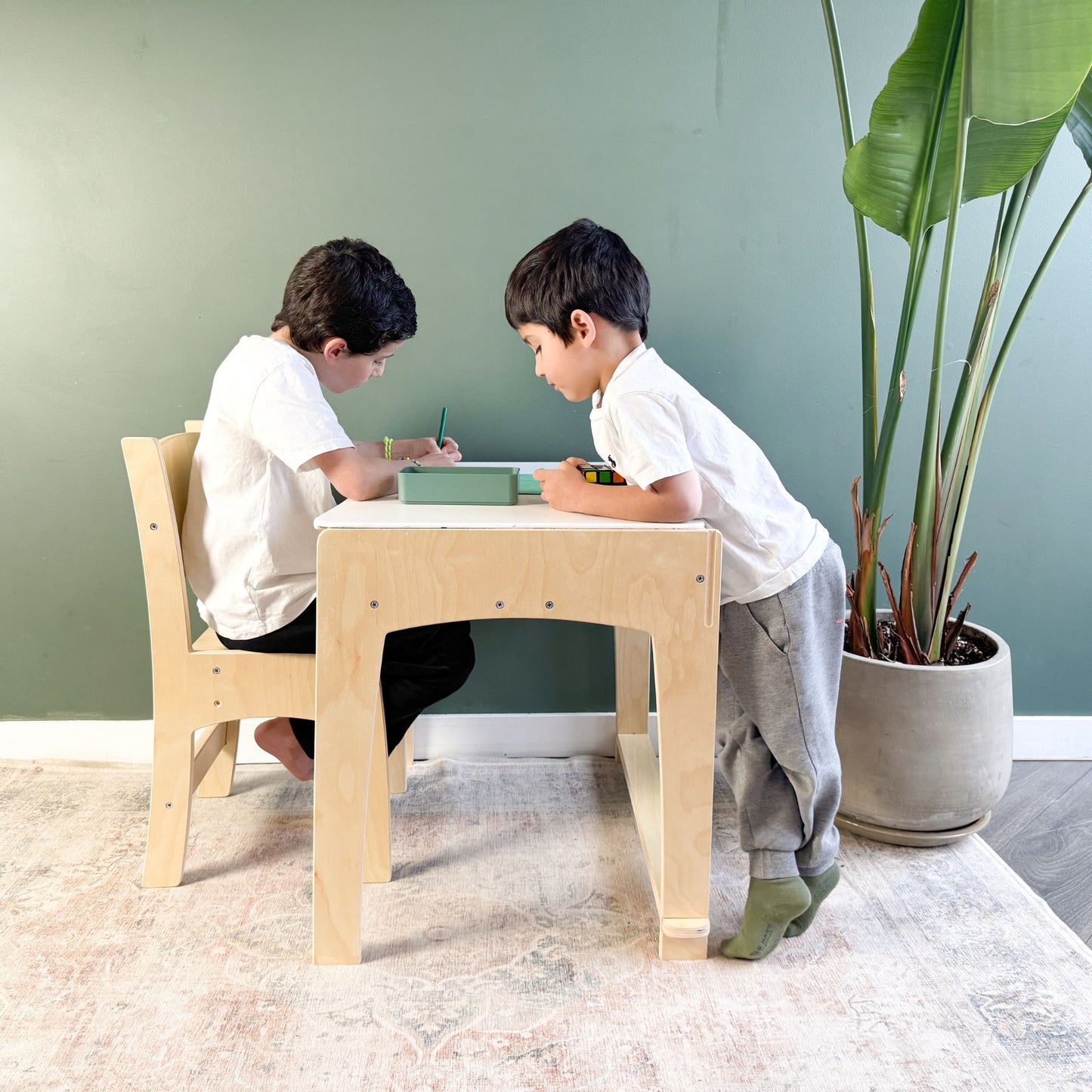 Two young children engaged in creative activities at a minimalist wooden desk and chair set. One child sits comfortably while drawing, while the other leans in with curiosity, demonstrating the inviting and collaborative nature of the space. The ergonomic furniture, set against a calming green wall with a large potted plant, fosters concentration, independence, and interaction in a Montessori-inspired learning environment