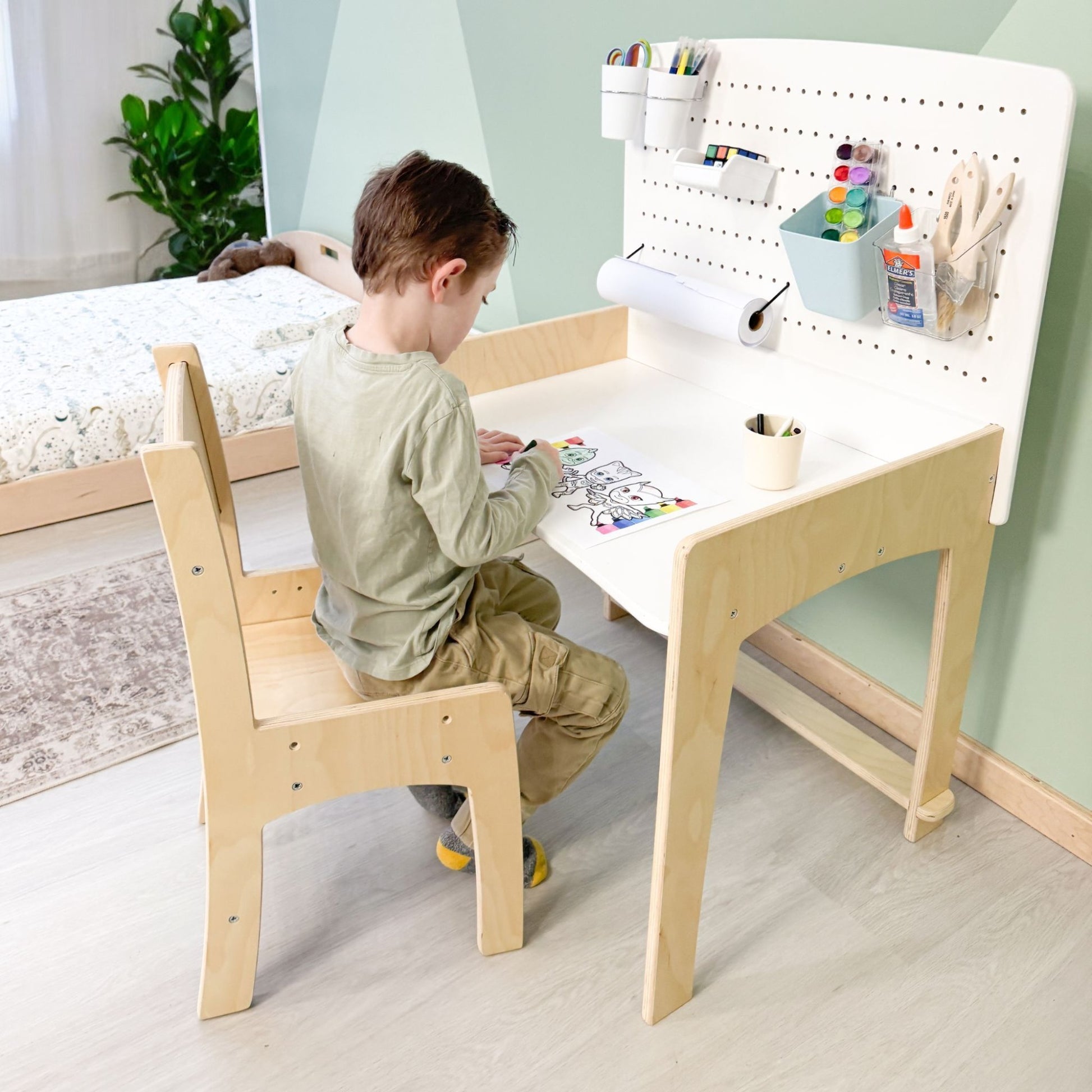 A young child sits at a Montessori-inspired wooden desk with an attached pegboard organizer, focused on coloring. The pegboard neatly holds art supplies, including paint, markers, brushes, and a paper roll, fostering creativity and organization. The minimalist desk and chair set, made of natural wood, is placed in a thoughtfully designed children's bedroom featuring a matching floor bed and soft, neutral tones, creating an inviting and functional learning space.