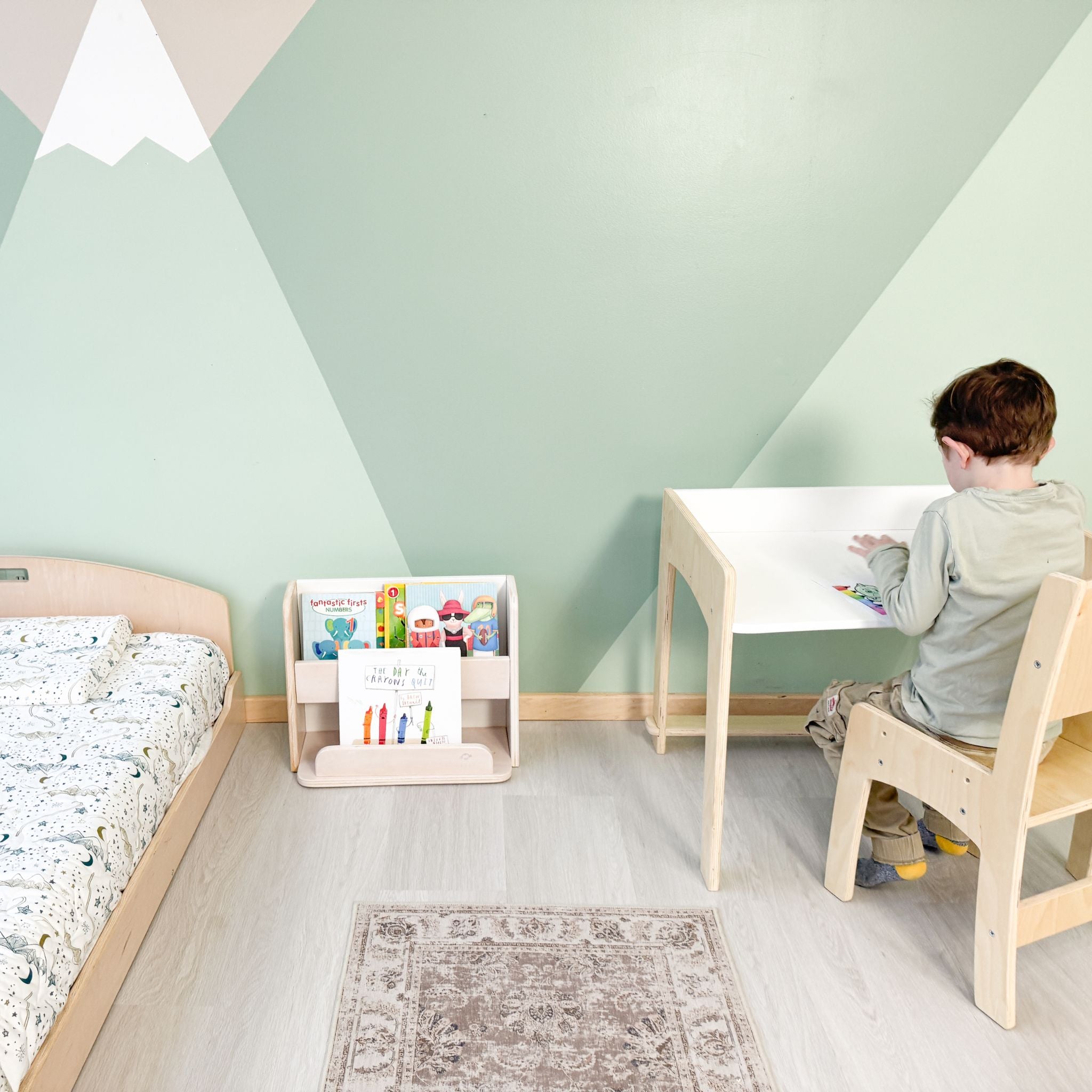 A cozy and organized Montessori-inspired children's room featuring a natural wood floor bed, a front-facing bookshelf filled with colorful books, and an adjustable wooden desk and chair set. A young child sits at the desk, engaged in an activity, while soft natural light enhances the calming green and geometric accent wall. The thoughtfully designed space fosters independence, creativity, and a love for learning.