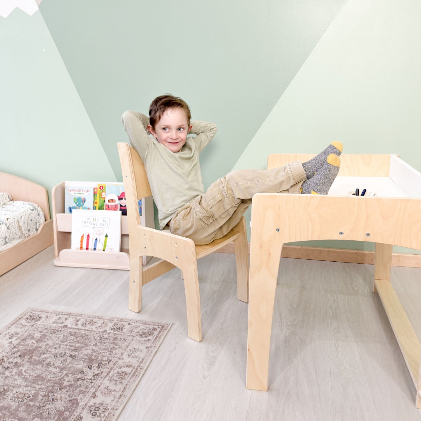 A confident young boy leans back in his adjustable Lumen Big Kid Desk and chair, exuding independence in his Montessori-inspired learning space. The sturdy wooden desk, designed to grow with him, provides the perfect setup for fostering focus, creativity, and self-reliance. In the background, a Montessori-style bookshelf encourages self-led learning, reinforcing an environment where children take charge of their development. Crafted in Canada 🇨🇦, this ergonomic workspace supports independence and confiden