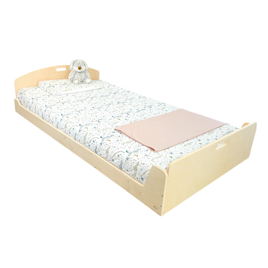 Low wooden twin bed with a curved headboard and side handles for easy movement. Designed for safe, independent sleep. Dressed in celestial-patterned bedding with a soft pink blanket and plush bunny. Ideal for Montessori-inspired bedrooms.