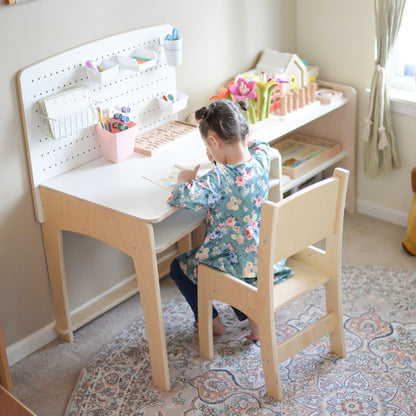 A young girl sits comfortably at the Lumen Big Kid Desk with an adjustable wooden chair, drawing and engaging in creative activities. The ergonomic children's desk features a spacious white melamine surface and a functional pegboard for organizing stationery, art supplies, and learning tools. A Montessori-inspired setup promoting independence and creativity, perfectly suited for kids aged 4.5 to 10+ years. Designed with high-quality birch wood and crafted in Canada.