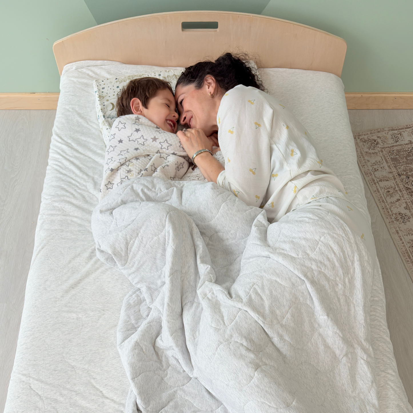 Close-up of a tender bedtime moment as a mother and child lie face-to-face under a soft quilt on a low wooden bed, capturing the emotional connection and sense of safety the design allows