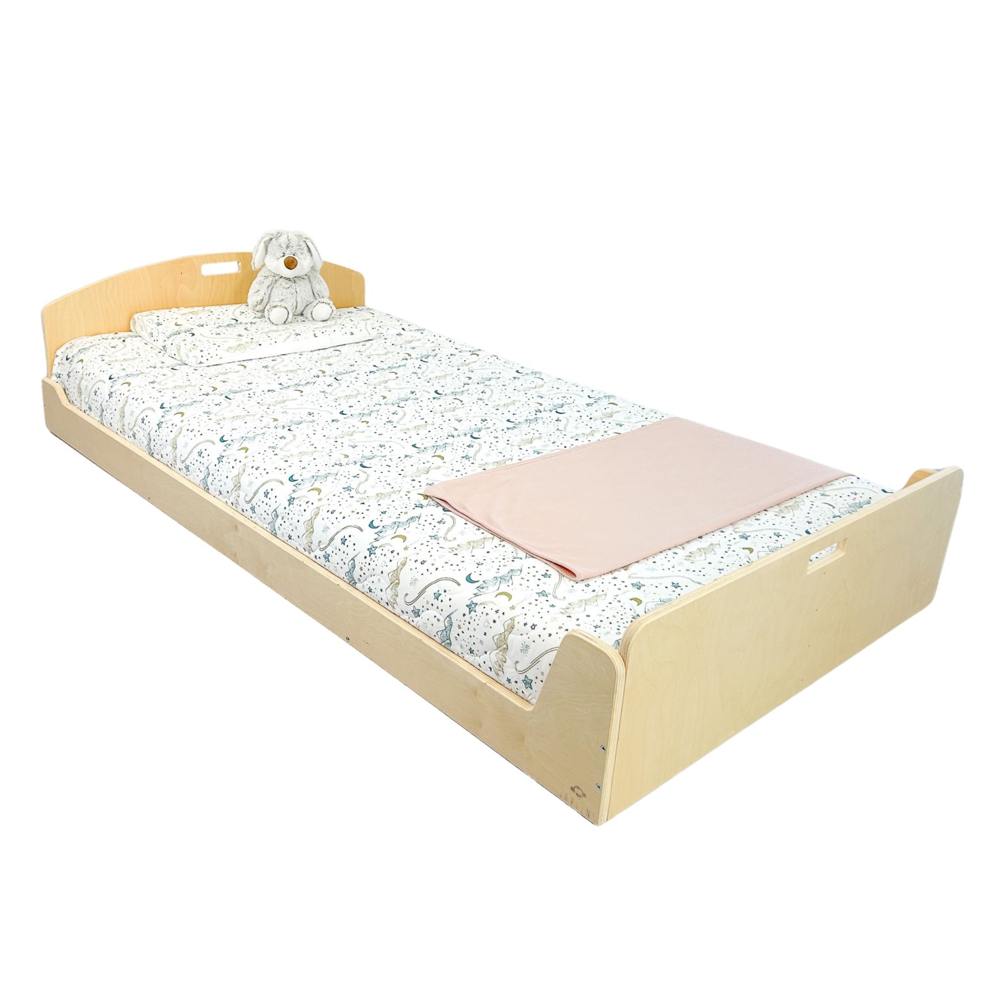 Low wooden twin bed with a curved headboard and side handles for easy movement. Designed for safe, independent sleep. Dressed in celestial-patterned bedding with a soft pink blanket and plush bunny. Ideal for Montessori-inspired bedrooms.