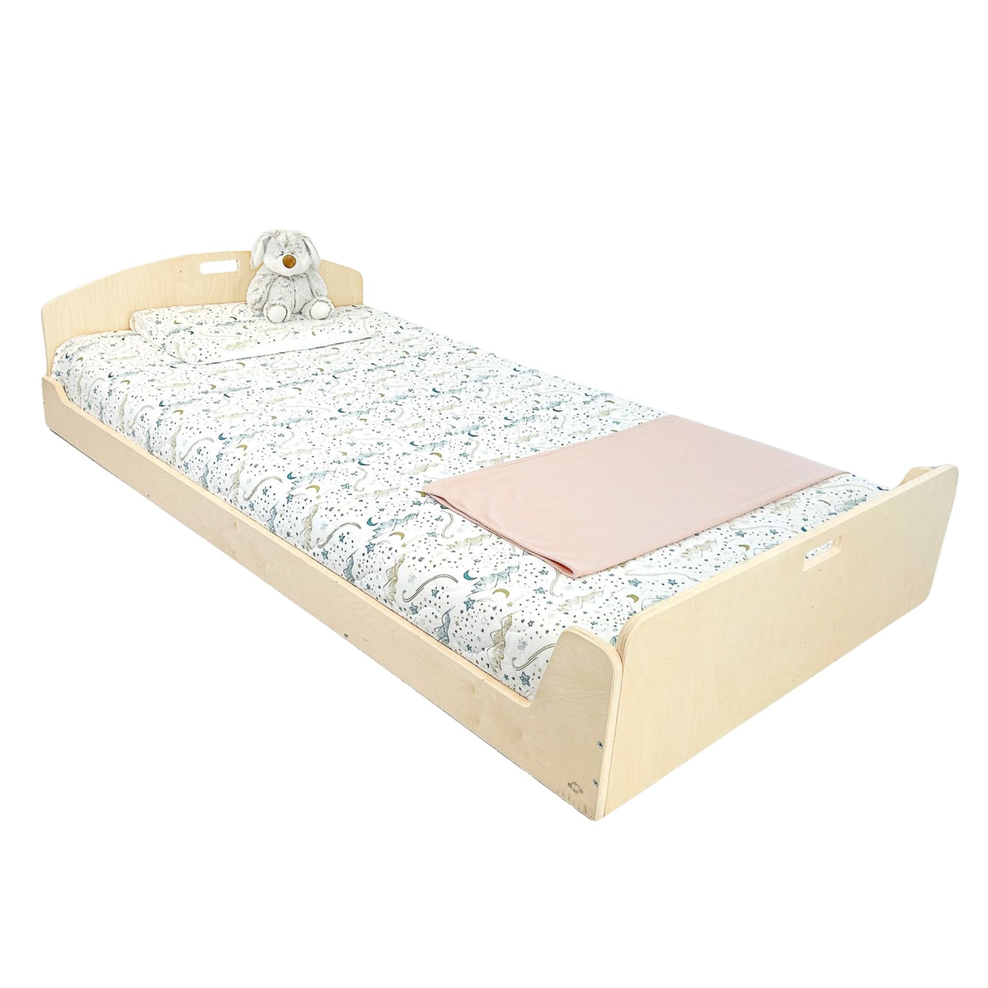 Low-profile wooden toddler bed with a natural finish, designed to promote independence in young children. Features an ergonomic curved headboard and built-in side handles for easy movement. Dressed with white bedding adorned with an animal-themed pattern in blue and beige, complemented by a gray plush bunny and a folded pink blanket at the foot of the bed. A safe and accessible sleeping solution for transitioning from a crib to a floor-level bed. Ideal for Montessori-inspired bedrooms or minimalist children