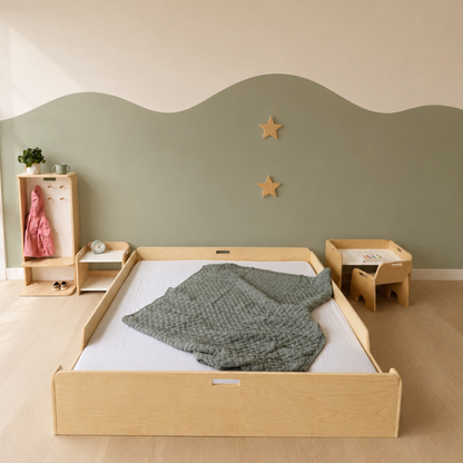 A light natural wood children's bed frame with a mattress and textured gray blanket, styled in a nursery with a sage green and cream wavy wall, wooden stars, and matching wooden storage furniture.