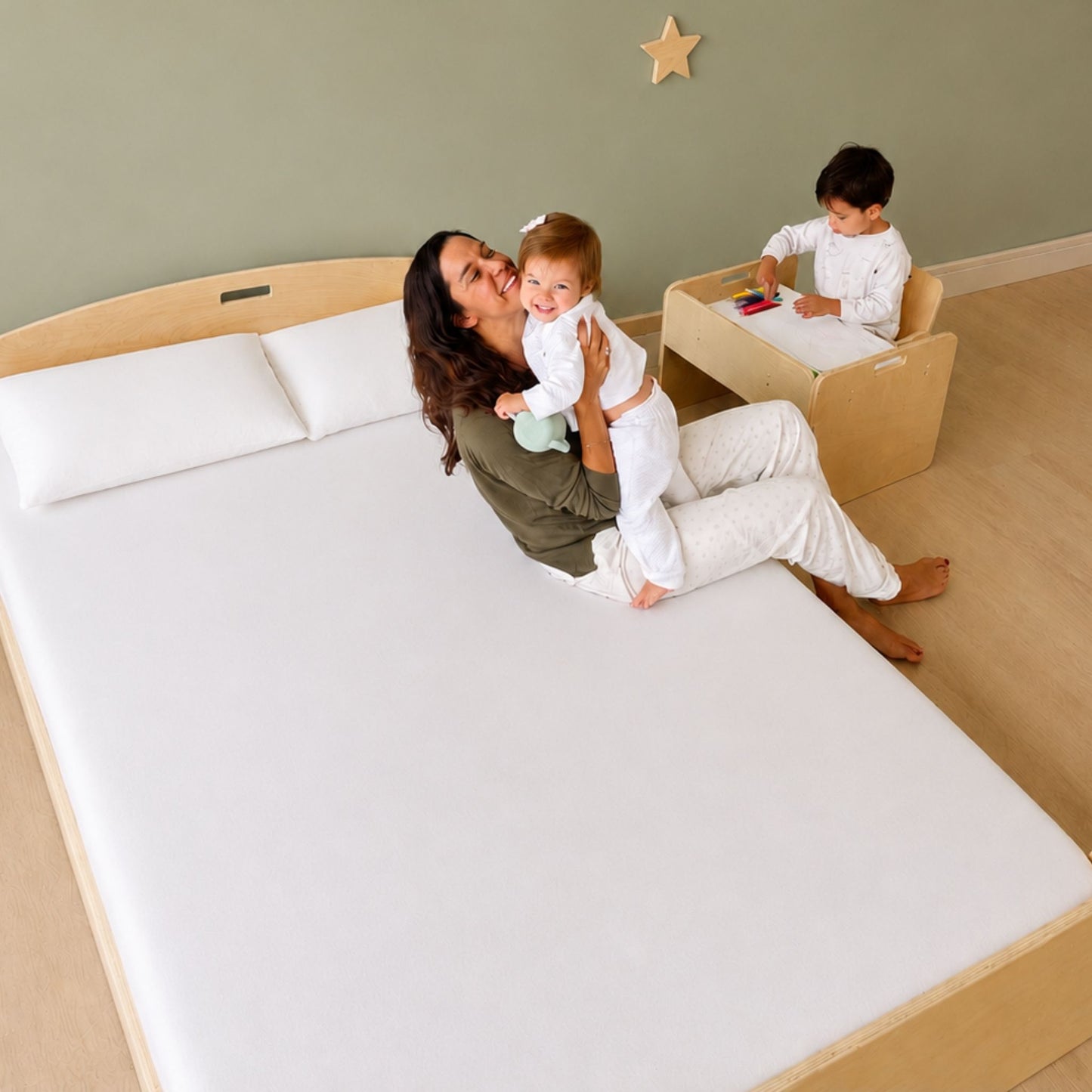 A woman sits on a large white mattress on a light wood bed frame in a nursery, holding a baby. In the background, a child plays at a small wooden activity table against a sage green wall.