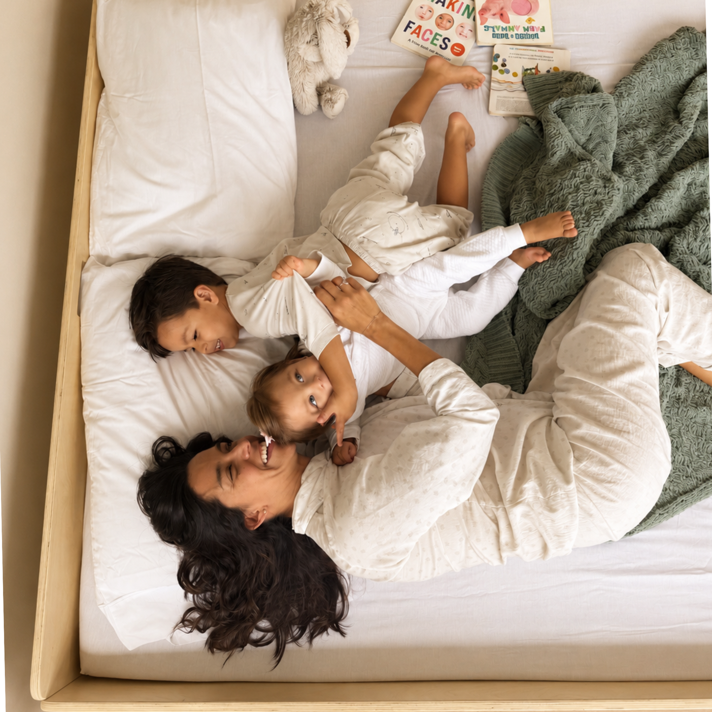 A family of three in matching white pajamas relaxing on a bed with white bedding, a green knit blanket, and children's books under a wooden headboard, viewed from above. The bed has wooden frames that keep toddlers in the bed at night.
