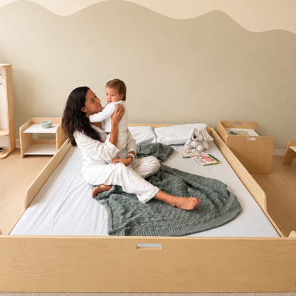 A woman in white pajamas holds a baby while sitting on a light wood Montessori toddler floor bed with a green knit blanket, in a softly lit nursery with matching wooden furniture.