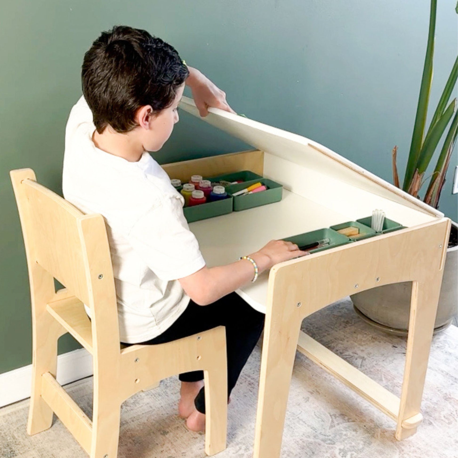 A child sitting at a natural wood adjustable desk, lifting the hinged tabletop to reveal a spacious built-in storage compartment. Inside, neatly organized art supplies, including paint, brushes, and pencils, are easily accessible. The ergonomic design promotes independence and organization, making it a perfect workspace for creative activities. The setup is complemented by a matching wooden chair and a calming green wall in the background.