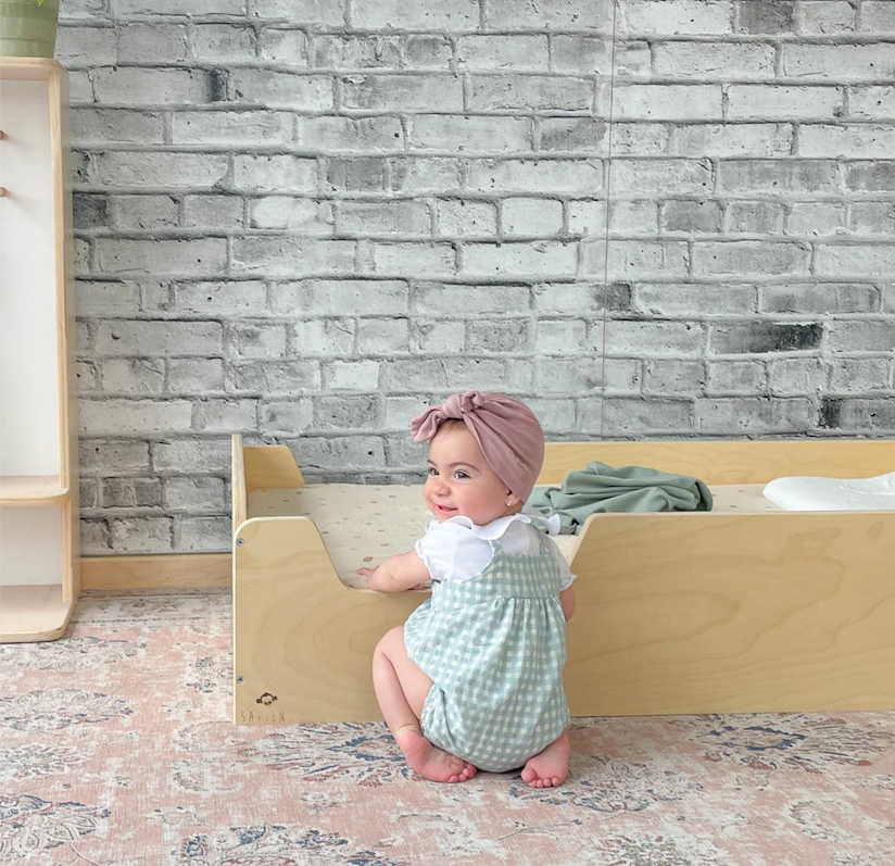 Baby sitting on a Montessori Floor Bed by Sapiens Child against a brick wall.
