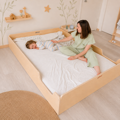 A woman and a child are sitting on a Montessori Floor bed from Sapiens Child in a child-friendly room with wooden furniture and decor.