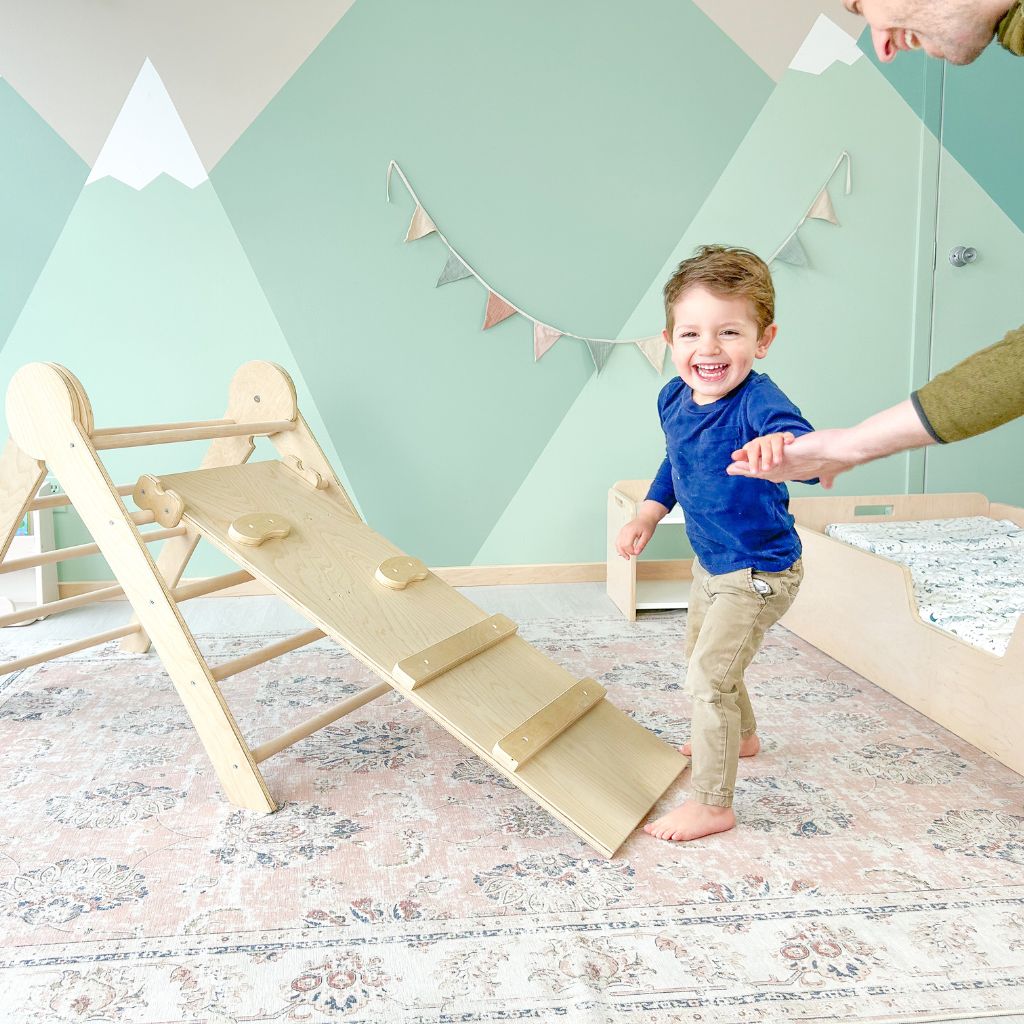 toddler smiling while holding dad's hand after climbing the wooden triangle pikler