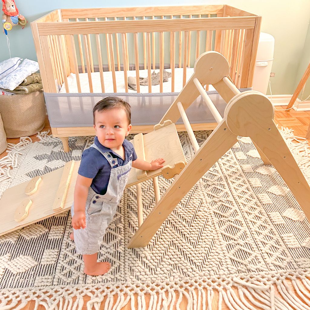 baby standing beside a wooden climber smiling and about to climb.