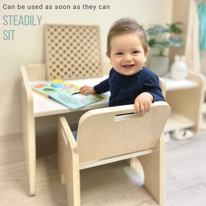 little one year old smiling boy with blue shirt sitting on the weaning chair. View is from behing but he is turning around and smiling. He was reading a book on top of the montessori activity table. The piece looks beautiful and steady.