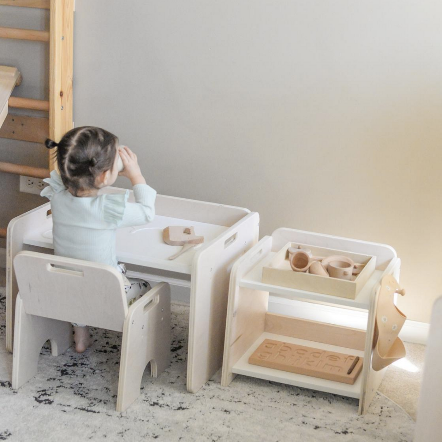 Toddler girl sitting on a wooden montessori chair in front of wooden montessori weaning table drinking from cup. Beside the table is the Papaya shelf by Sapiens Child . The shelf has a wooden tray with wooden toys and a hook on the side that holds a beige bib.